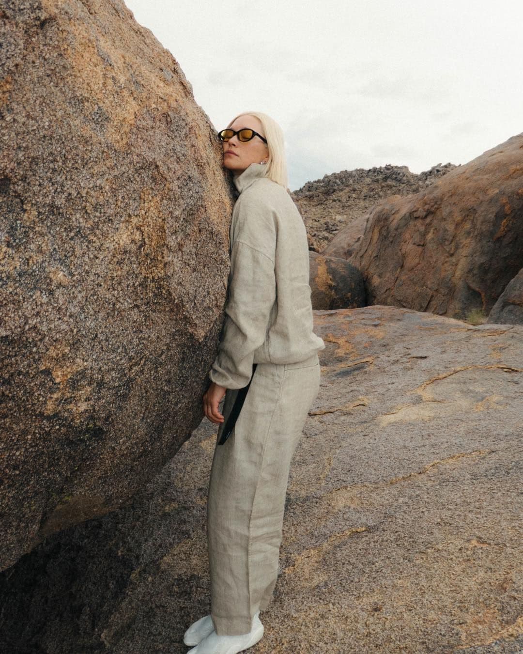 A blonde woman in a light linen outfit and yellow sunglasses leans against a large rock in a rocky landscape.
