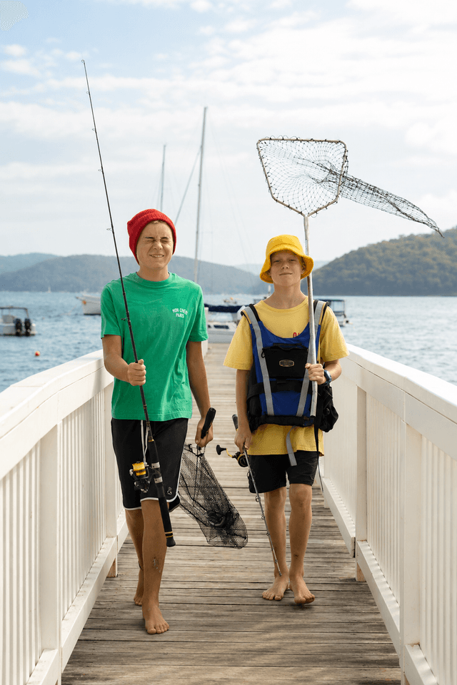 Two boys walk barefoot on a pier, carrying fishing rods and nets, with boats and hills in the background.