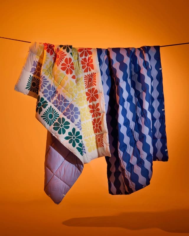 Two quilted blankets hang on a line against an orange background; one features a colorful floral pattern, the other a blue and white wavy design.