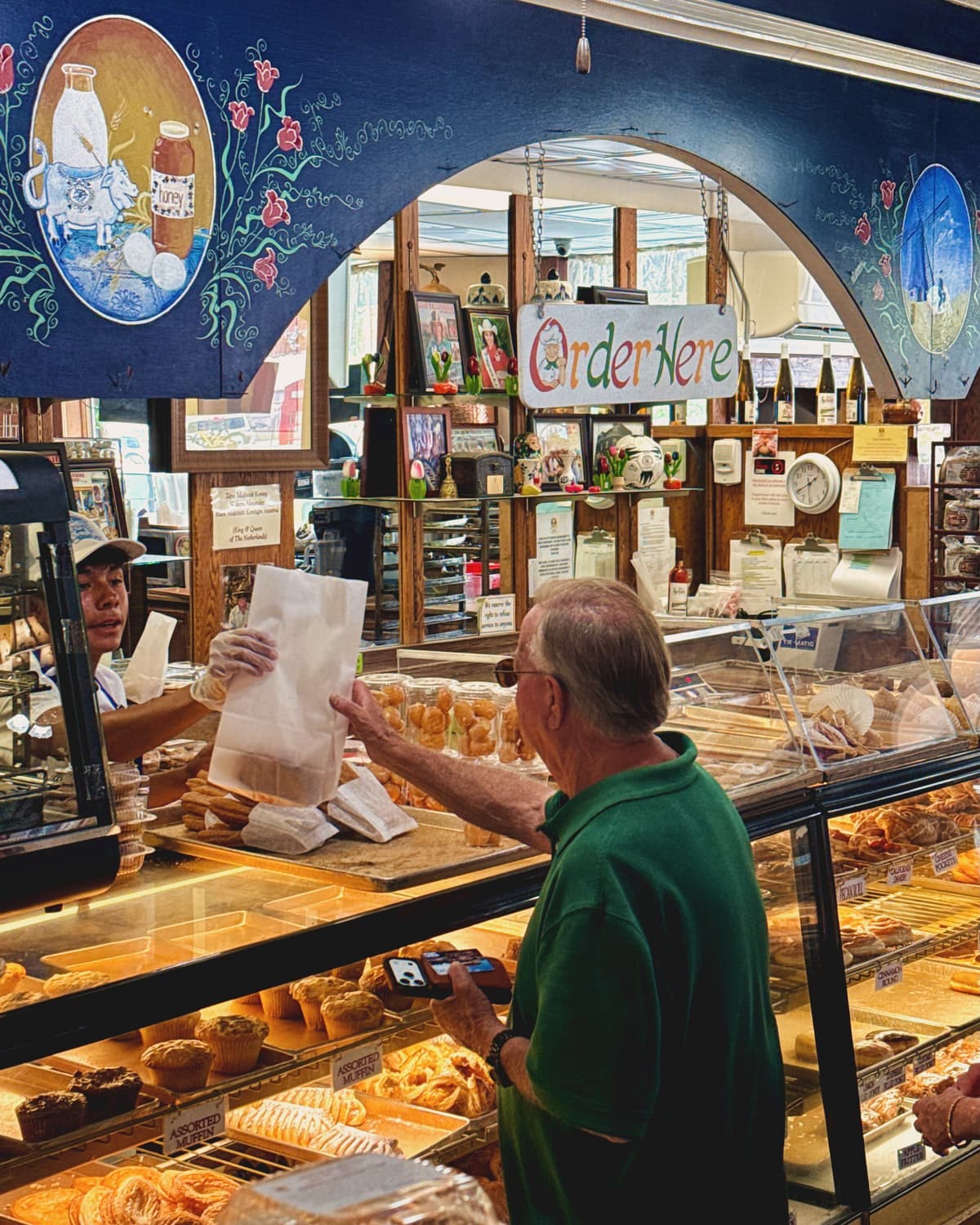 An employee hands a bag of baked goods to a customer in a vibrant bakery with pastries on display and decorative murals.