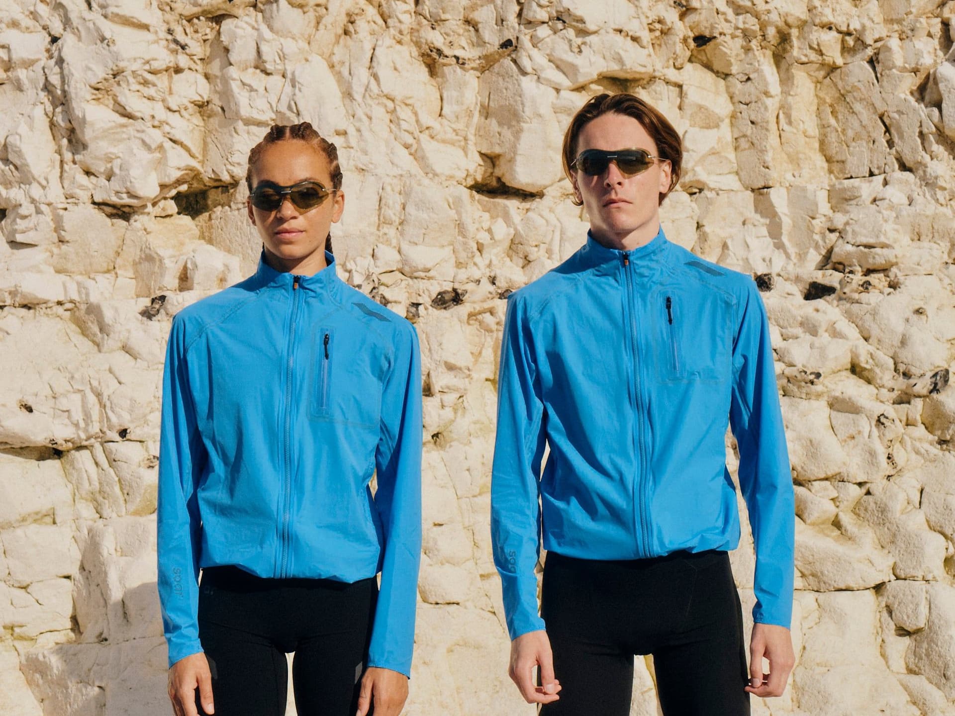 Two athletes in blue jackets, black athletic wear, and sunglasses running outdoors.