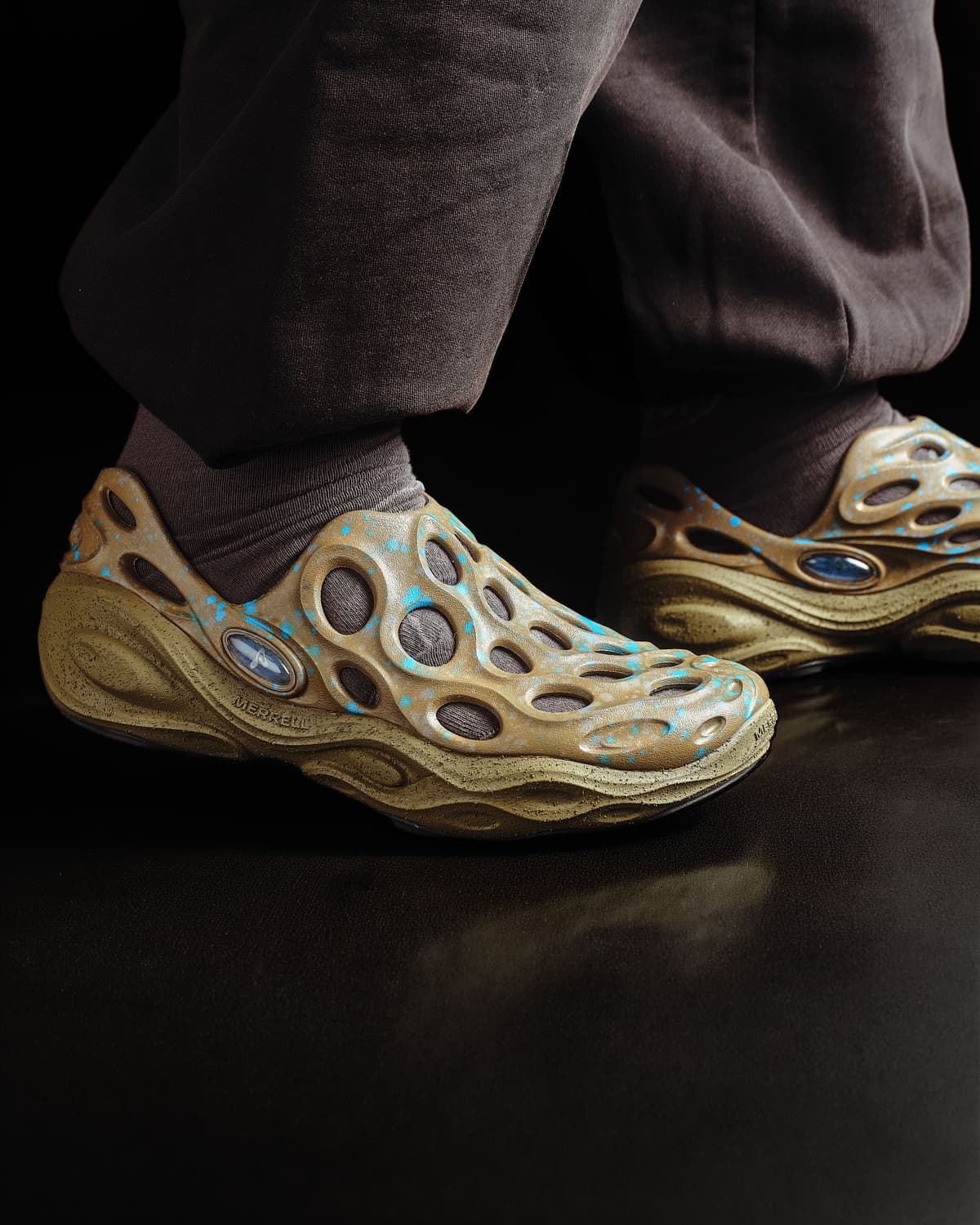 Beige, perforated shoes with blue splatters worn by a person in brown pants.
