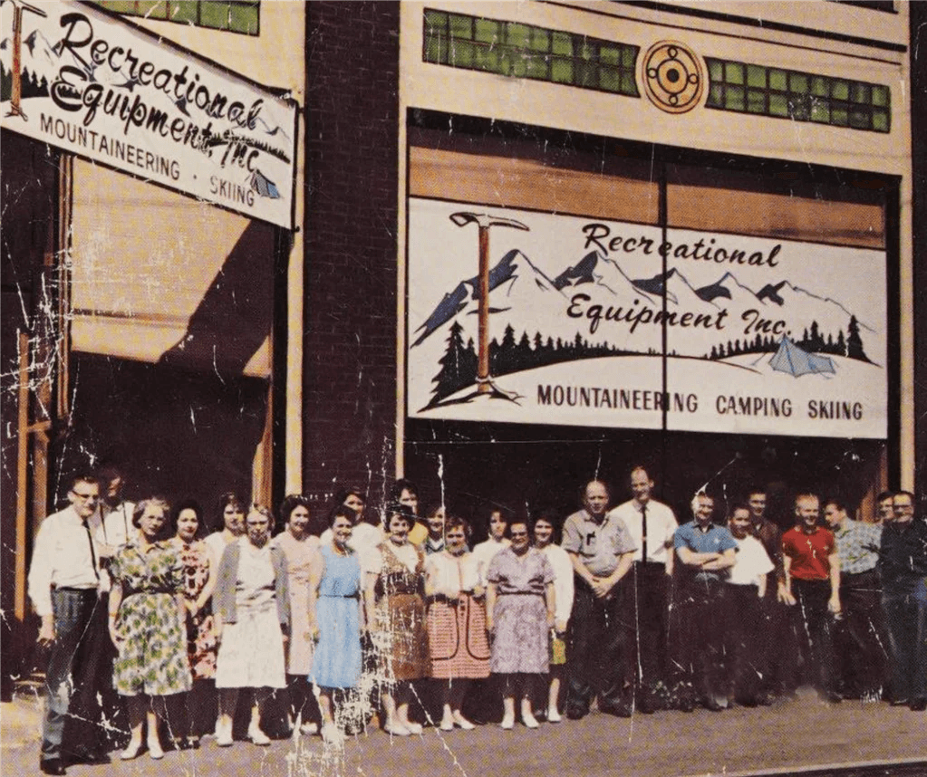 A group of employees stands in front of a vintage Recreational Equipment Inc. store.