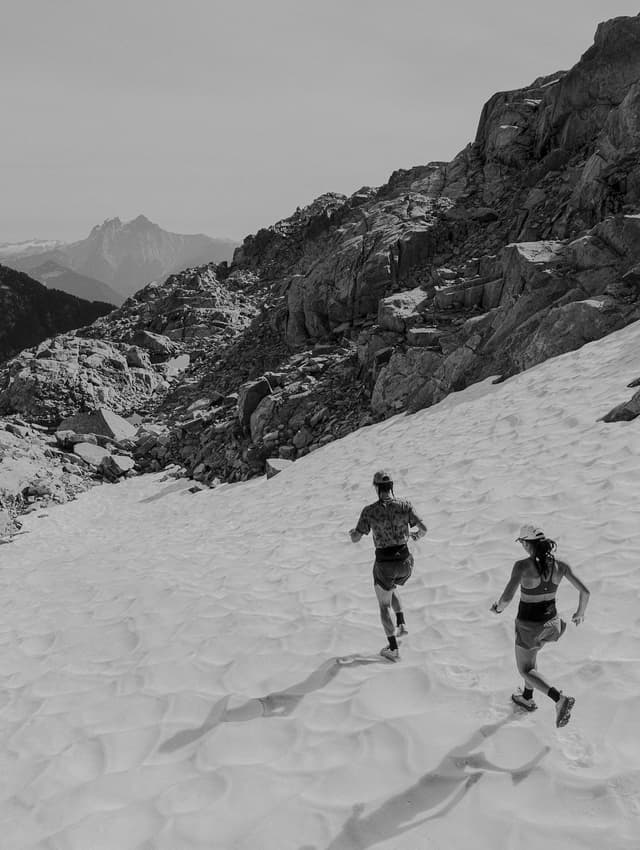 Black and white photo of two people trail running uphill on a snow-covered mountain slope.