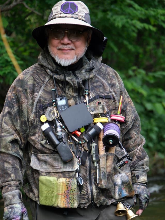 A smiling man with a white beard and glasses wears a camouflage fishing jacket covered in various gear and a bucket hat with a purple peace sign.