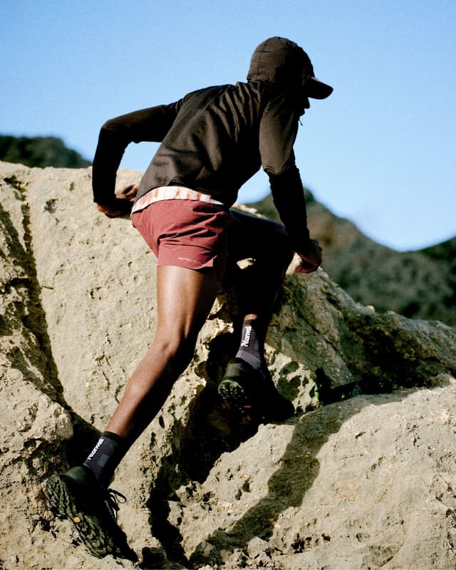 A man in a dark hoodie and red shorts scrambles over sunlit rocks.