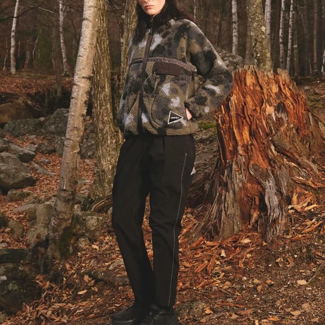 A person in a green and grey camo fleece jacket, black pants, and black shoes stands in a forest with trees and a large stump.