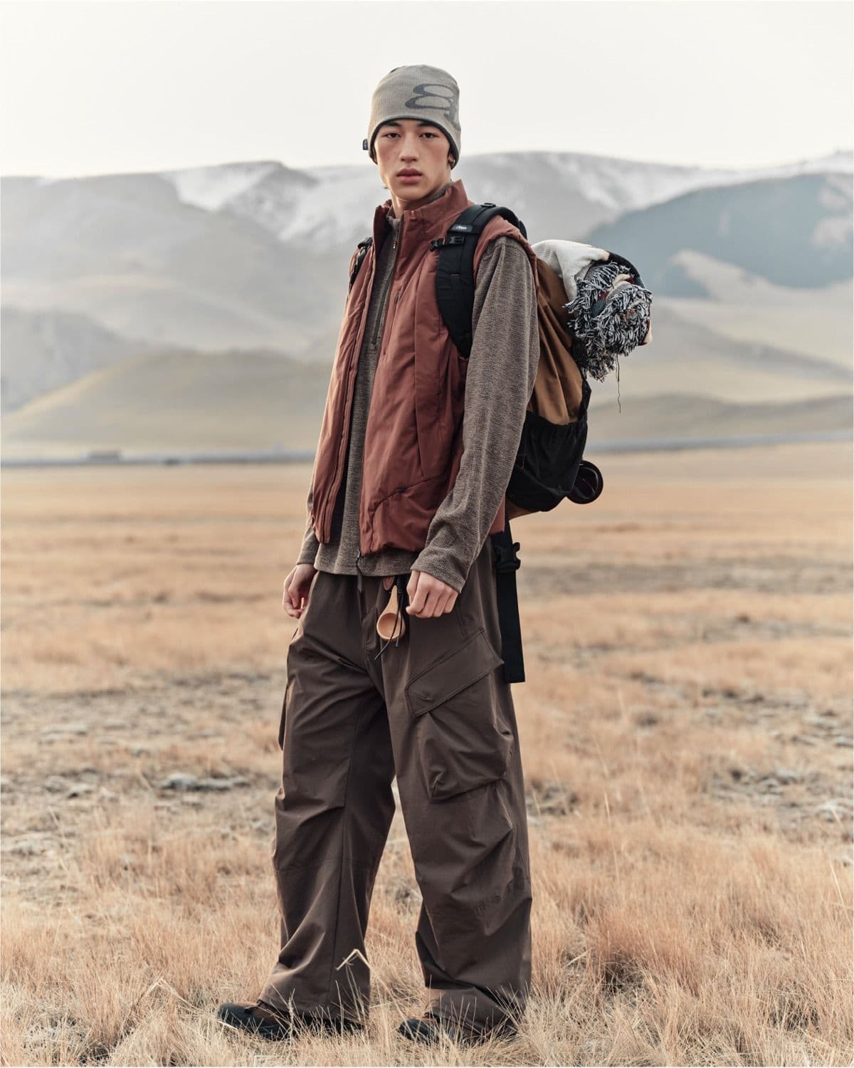 A person in outdoor gear, including a beanie, rust vest, and cargo pants, stands in a dry field with mountains in the background.