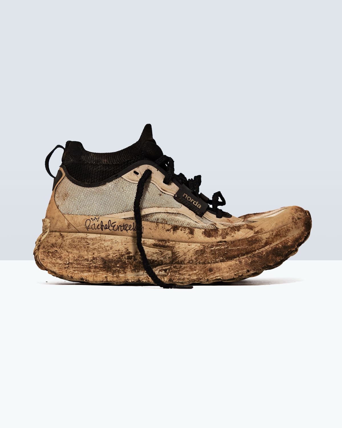 A mud-caked Norda trail running shoe with black accents and "Rachel Kunkel" handwritten on the side.