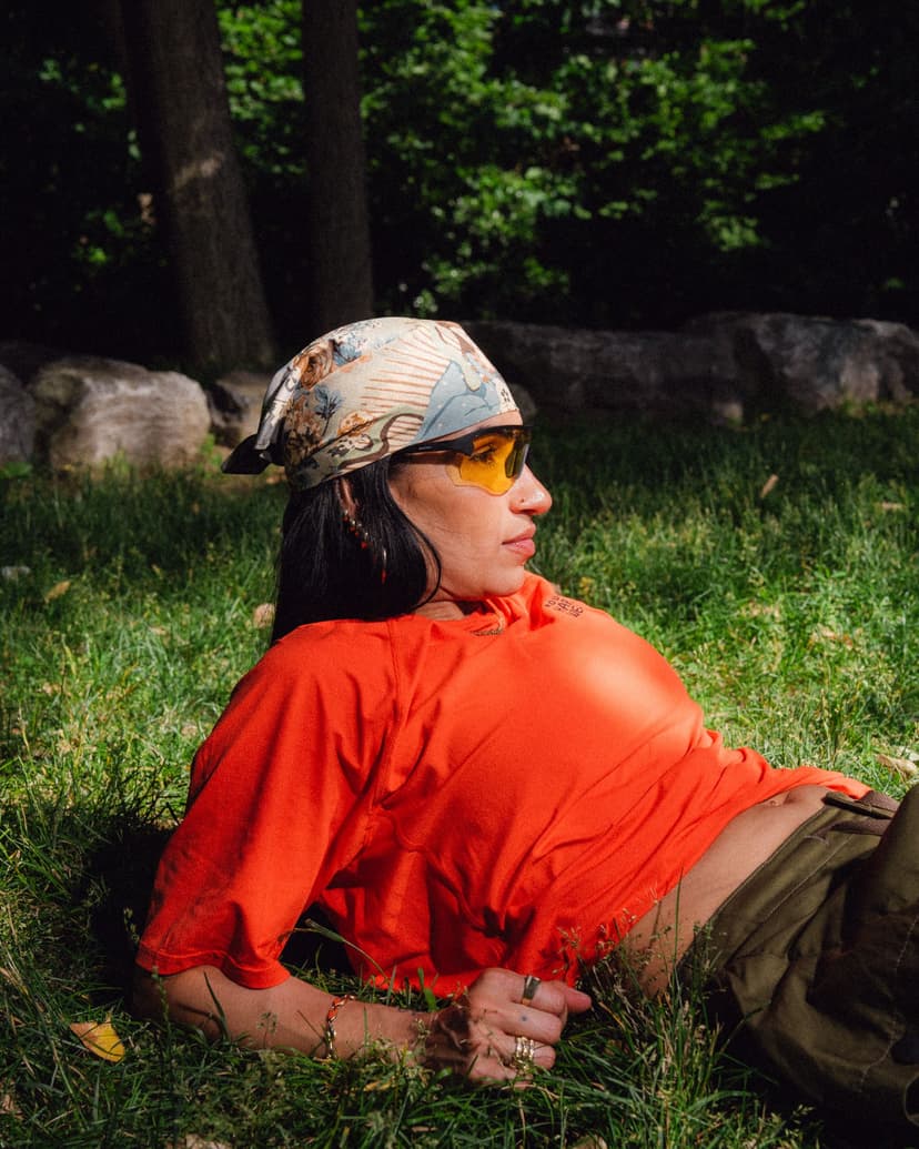 A person wearing a patterned bandana, yellow sunglasses, and an orange shirt reclines on their side in green grass.