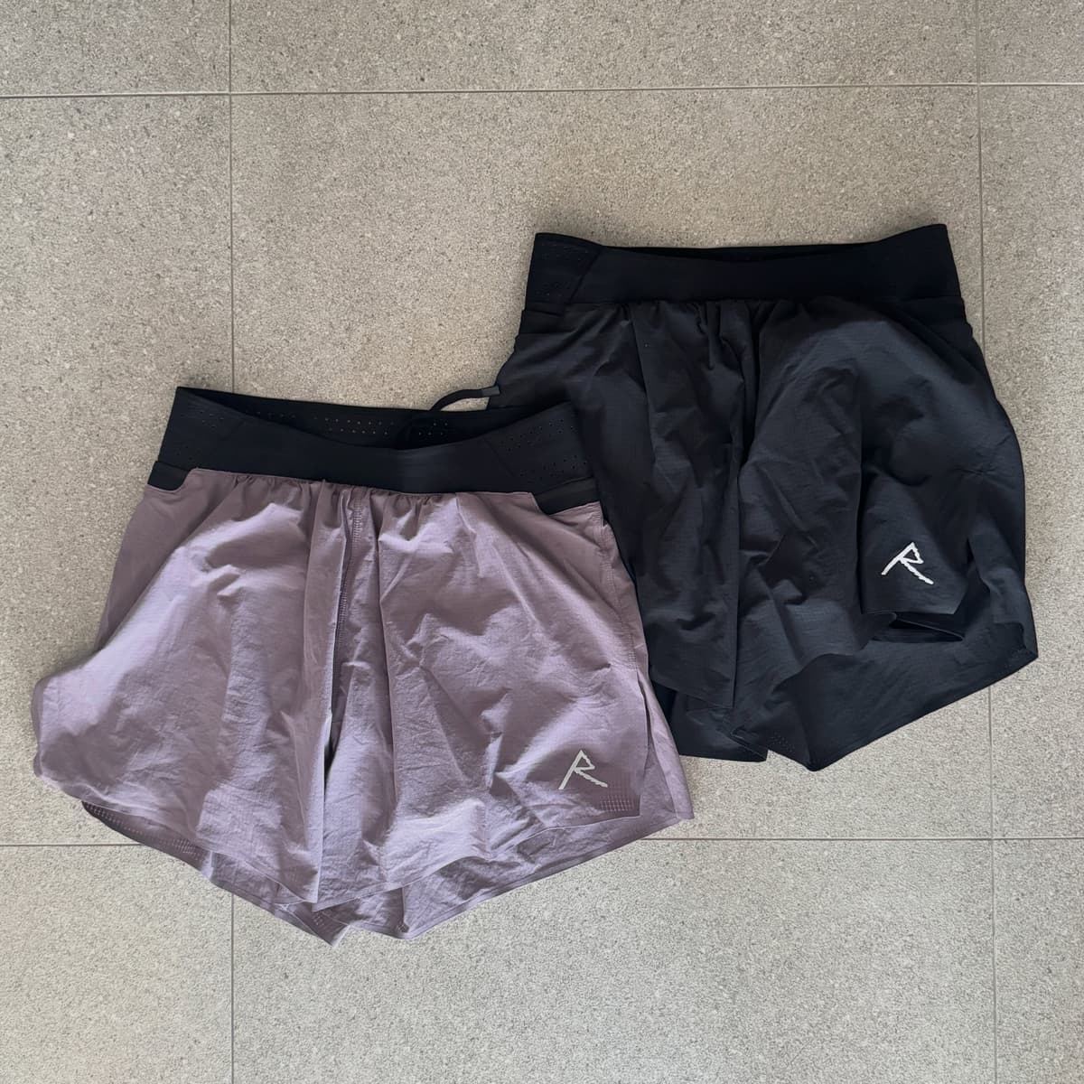 Two pairs of athletic shorts, one light purple and one black, each with a white 'R' logo, laid on a tiled surface.