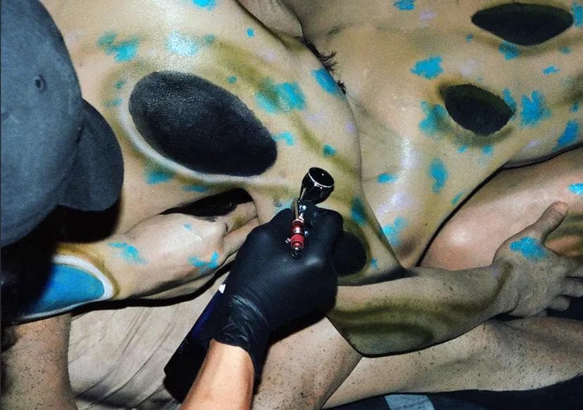 Close-up of a person's torso being airbrushed with blue, black, and tan body paint.