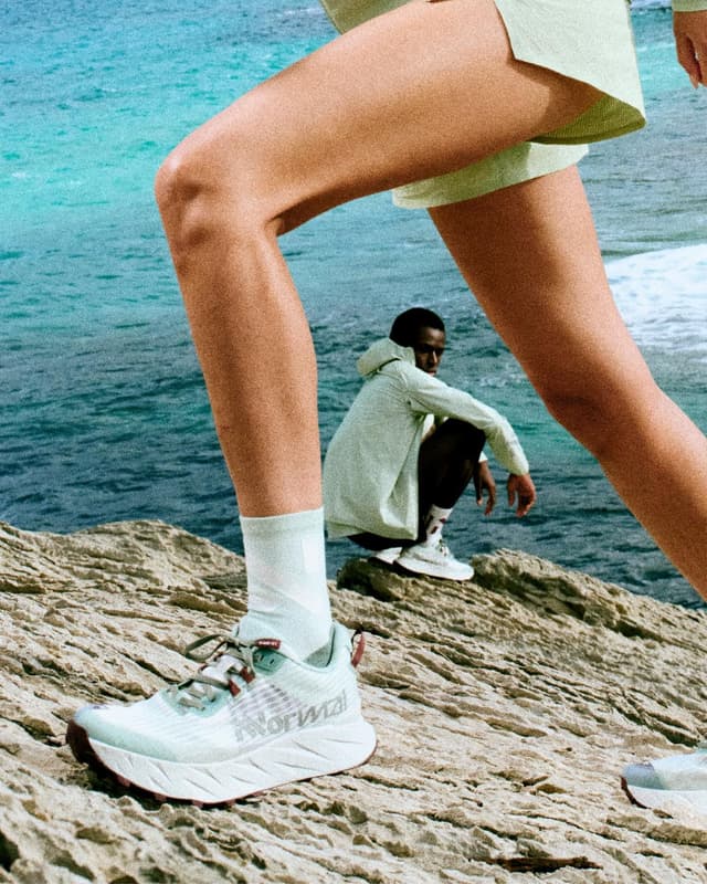 Two people on a rocky coast: one in the foreground steps forward wearing light green activewear and Normal running shoes, and another crouches by the ocean.