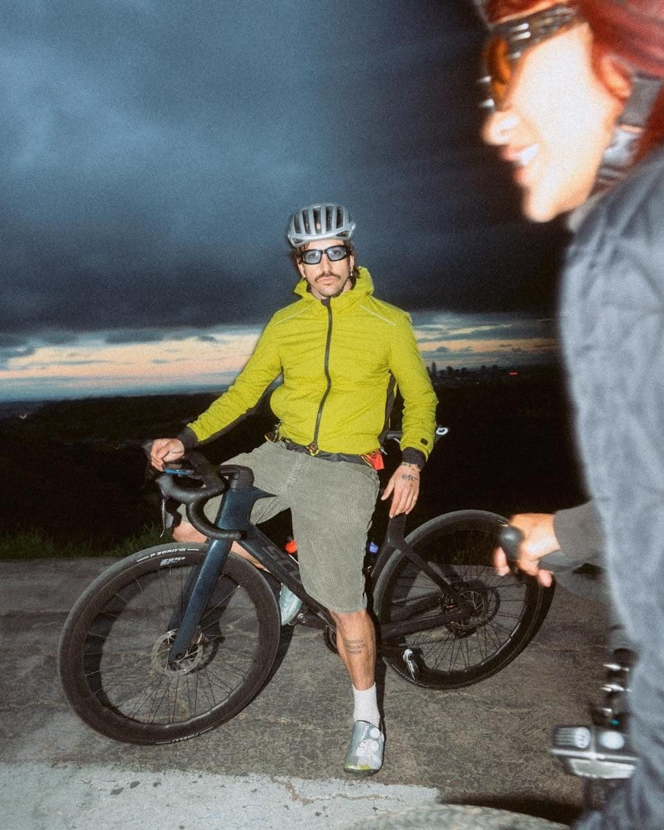 A cyclist in a lime green jacket and helmet stands with his bike under a dark, cloudy sky, with another cyclist blurred in the foreground.