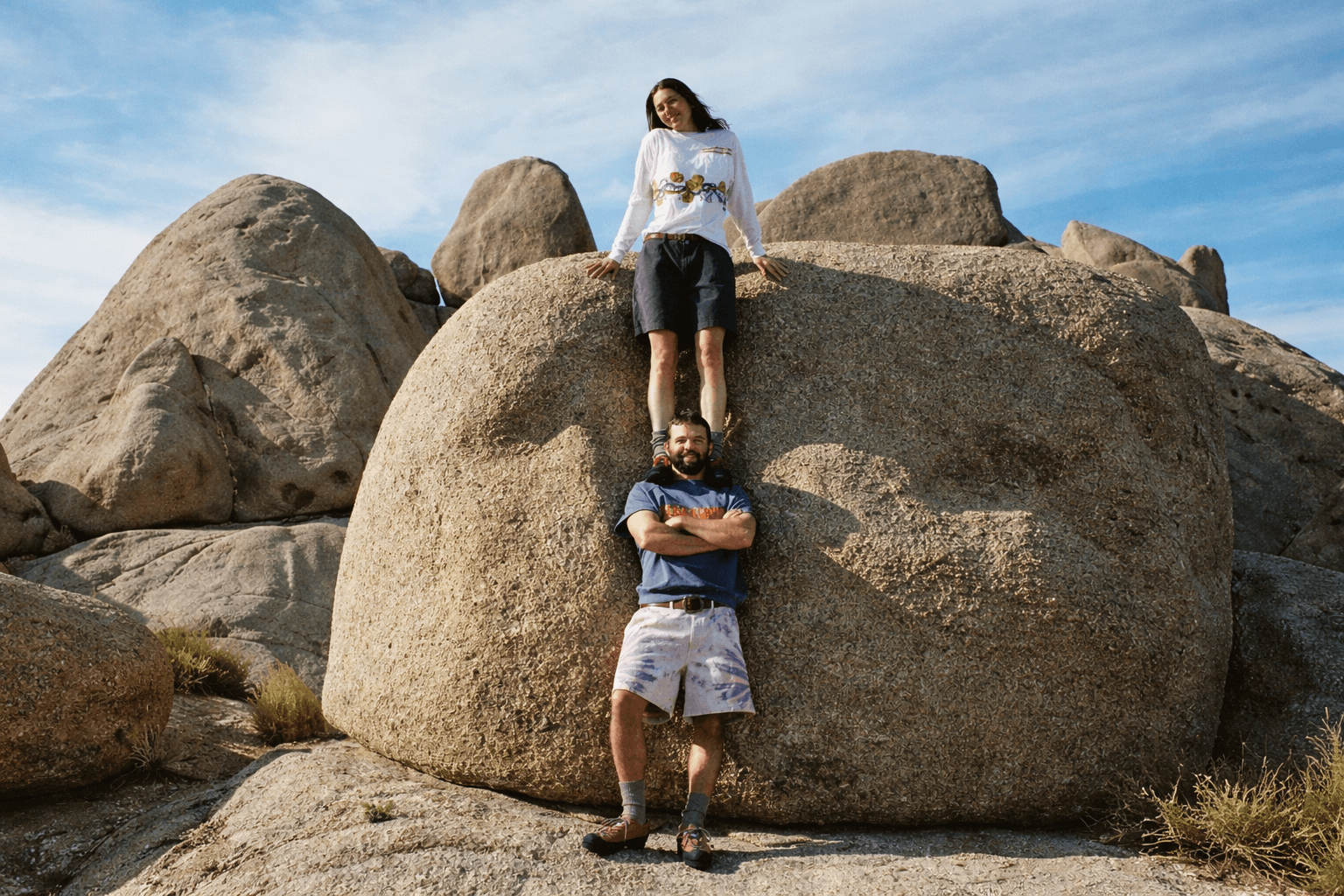 A woman stands on a man's shoulders among large boulders under a blue sky.