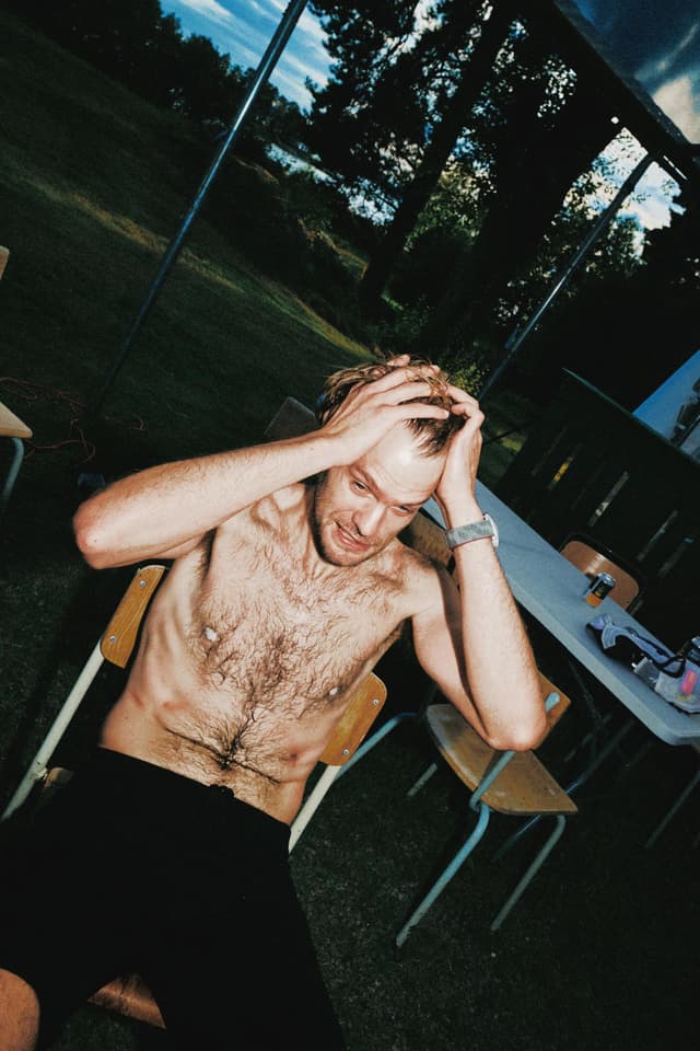 A shirtless man with blonde hair sits on a chair outdoors, hands on his head with a pained expression, against a background of grass, trees, and a lake.