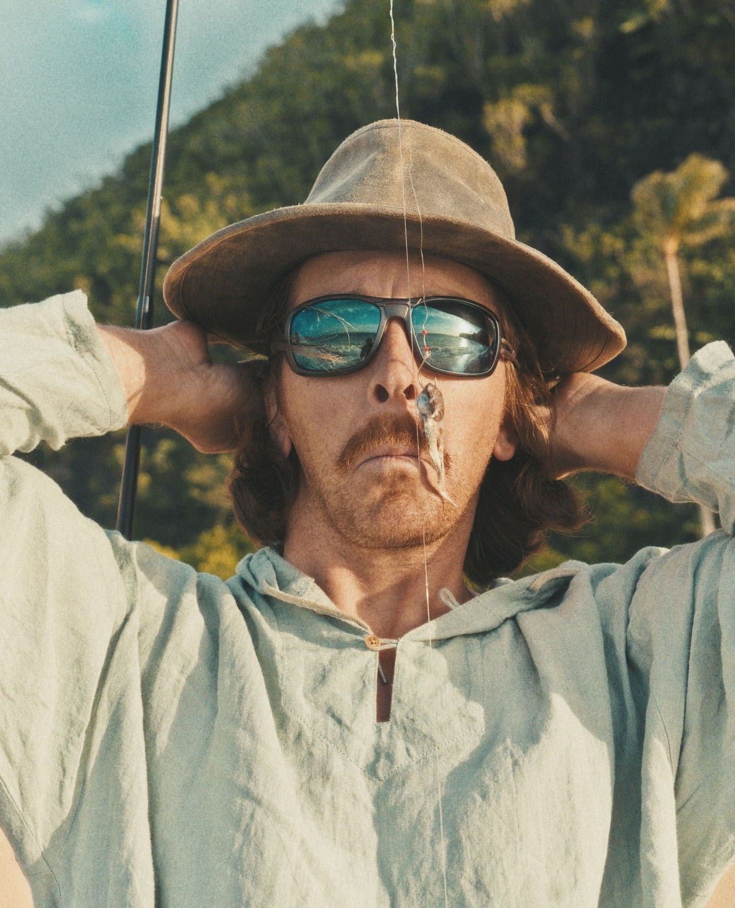 A man in a hat and sunglasses with a small fish on a fishing hook dangling in front of his face, hands behind his head.