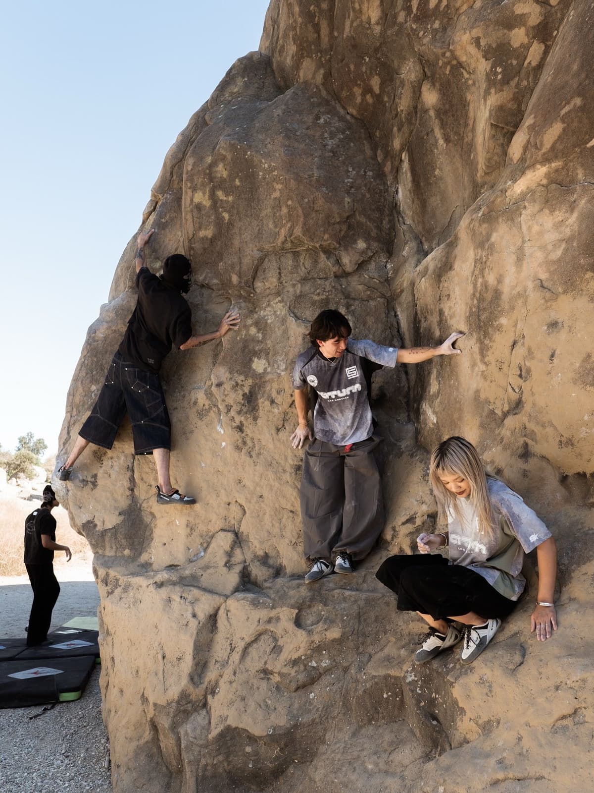 Four people bouldering on a large outdoor rock, with crash pads below.