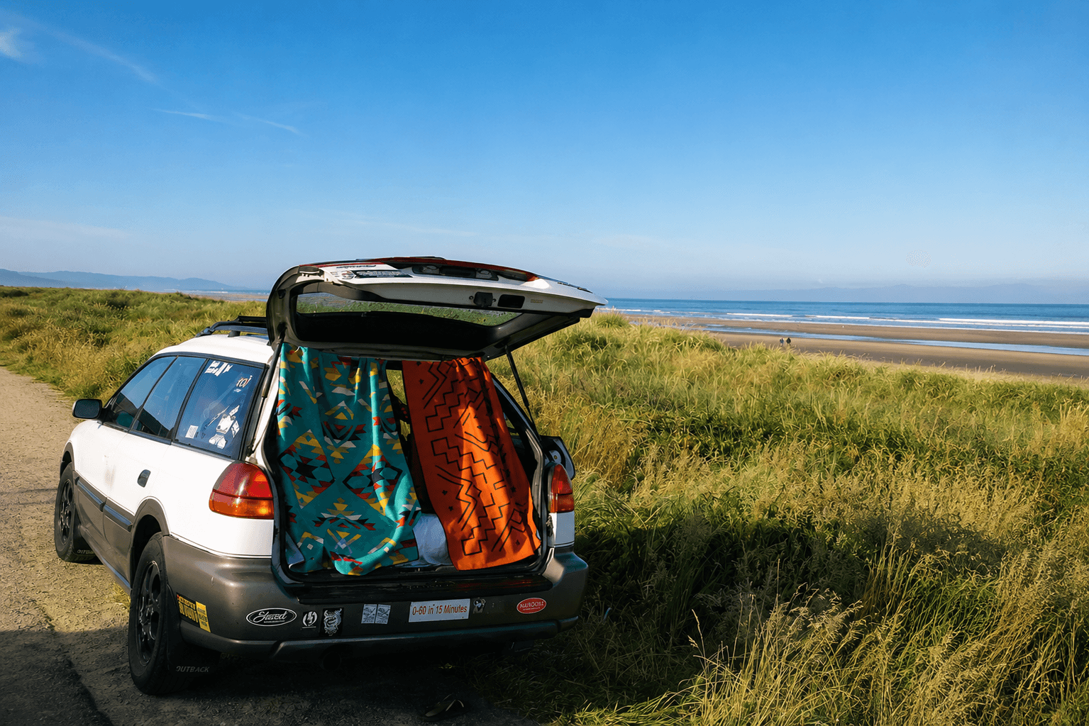 A white Subaru wagon with two colorful blankets hanging from its open trunk, parked by a grassy dune overlooking a sandy beach and ocean.