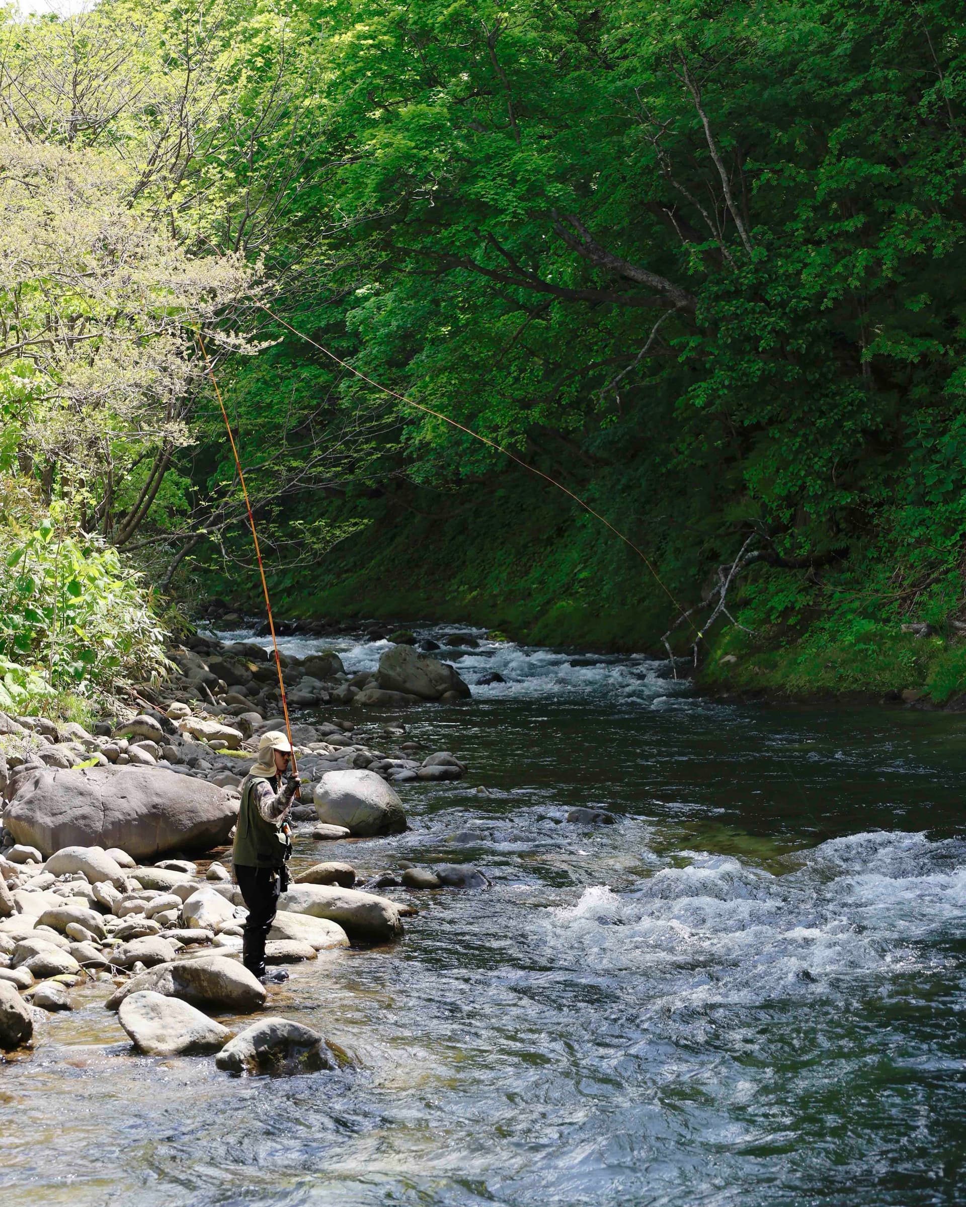 A person fly fishing in a rocky river surrounded by lush green trees.