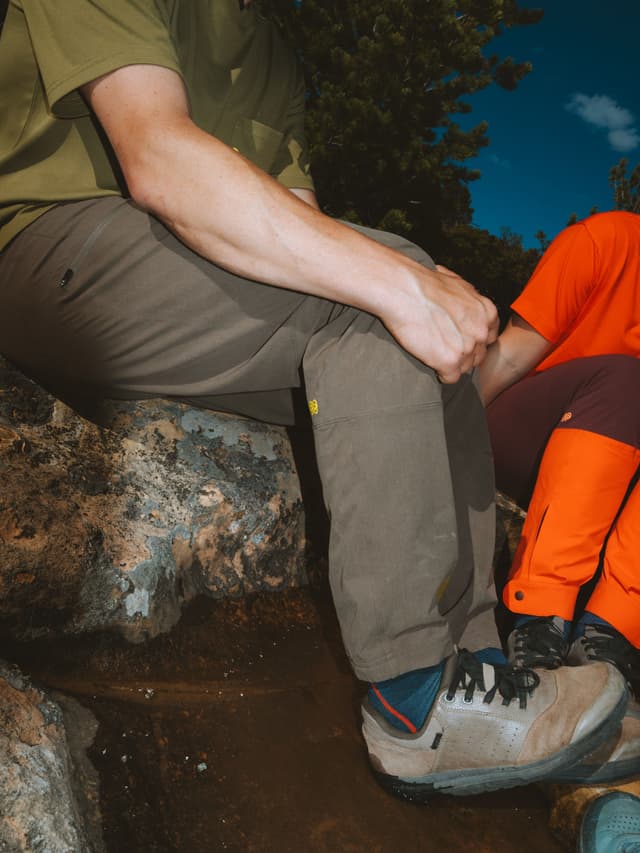 Two people, one in green and one in orange outdoor clothing, sit on rocks in a natural setting.