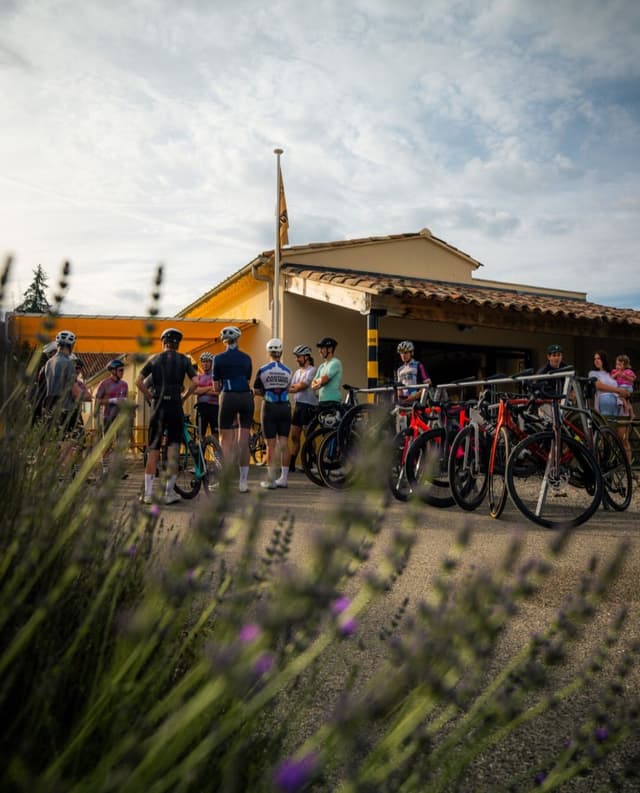 A group of cyclists with bikes stands in front of a building with a yellow awning, with lavender in the foreground.