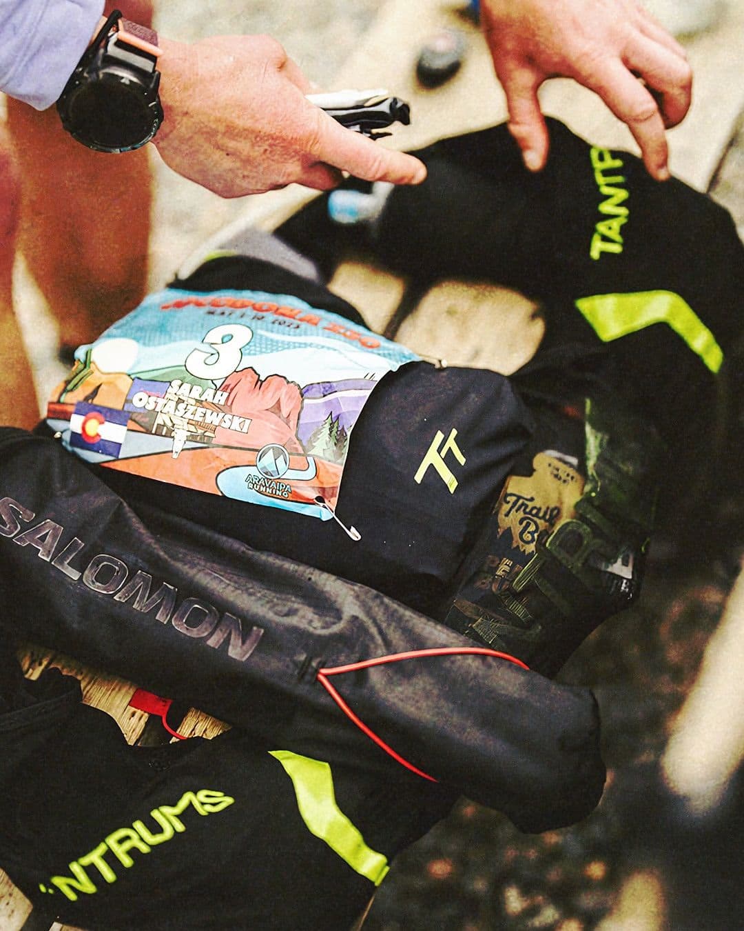 Overhead view of hands arranging trail running gear, including a race bib for Sarah Ostaszewski (number 3) and a Salomon pack.