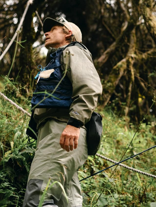 Man in fishing vest and waders looks up in a dense forest.