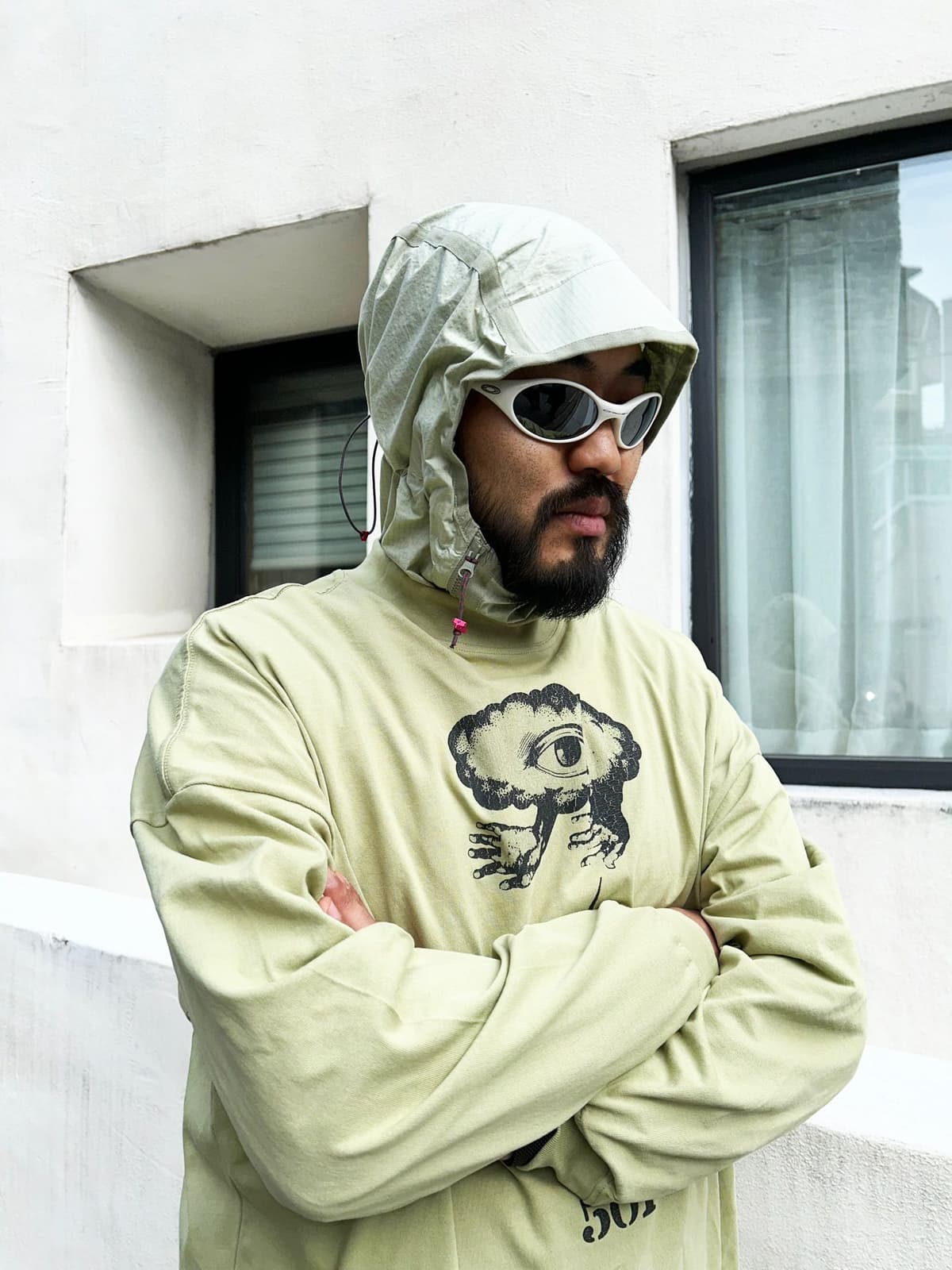 A bearded man in a light green hooded top with an eye-cloud graphic, white sunglasses, and crossed arms.