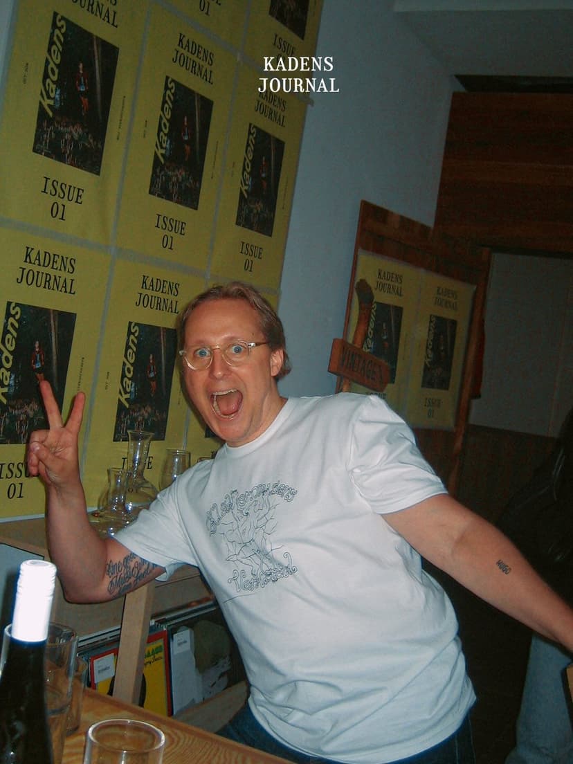 A man in glasses makes a peace sign and shouts excitedly in front of a wall covered with "Kadens Journal Issue 01" posters.