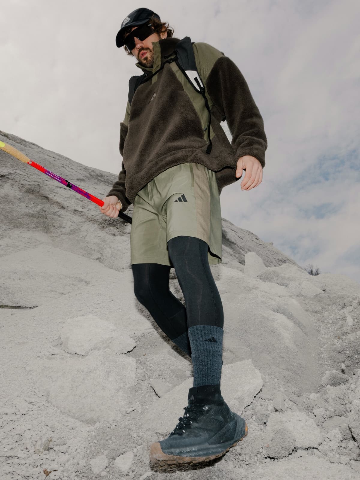 A person in a brown fleece, olive shorts over black leggings, and black hiking boots descends a rocky terrain, holding a colorful hiking pole.