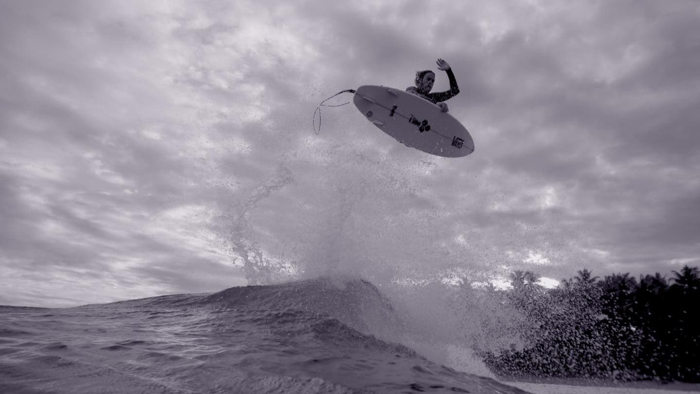 Black and white image of a surfer airborne above a breaking wave, with palm trees visible on the shore.