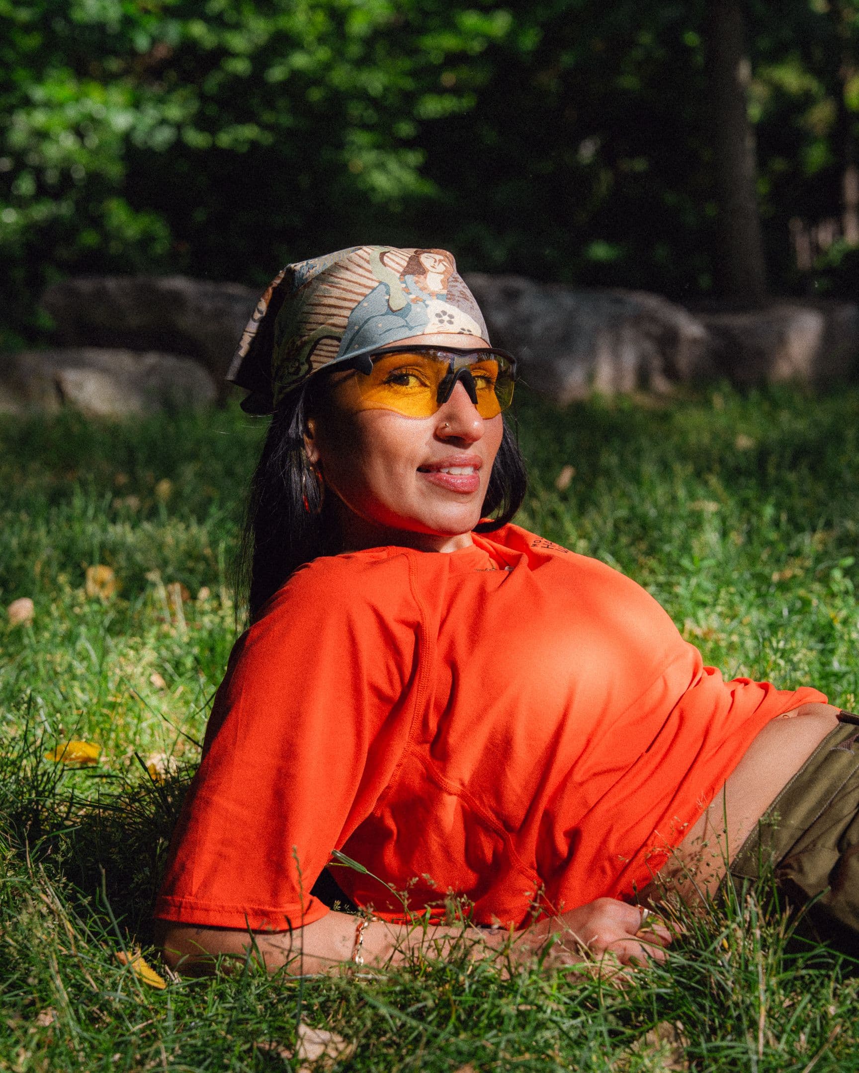 A smiling woman in an orange shirt, yellow sunglasses, and a patterned bandana lies on green grass.