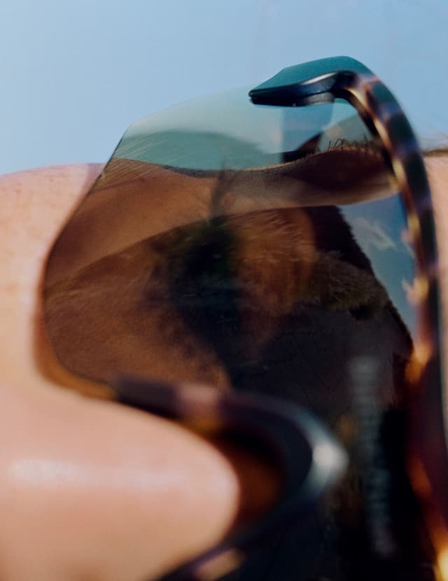 Close-up of a person wearing tortoise-shell sunglasses reflecting a sandy landscape and blue sky.