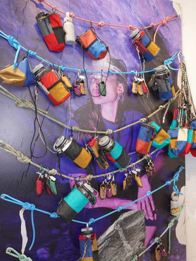 A wall display of climbing ropes strung over a large poster, adorned with colorful water bottle holders and miniature climbing shoe keychains.