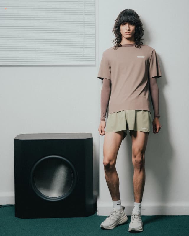 A person wearing a brown t-shirt, green shorts, and white sneakers stands beside a large black speaker.
