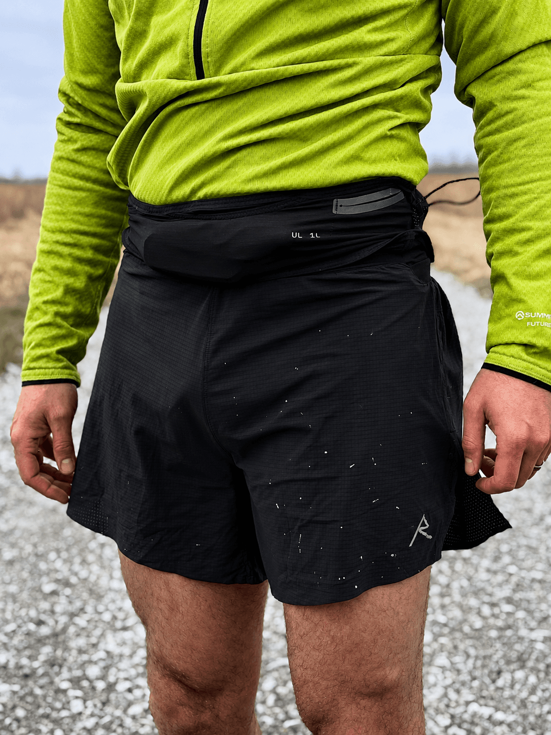 A person in a bright green shirt, black running shorts, and a black UL 1L running belt, standing on a gravel path.
