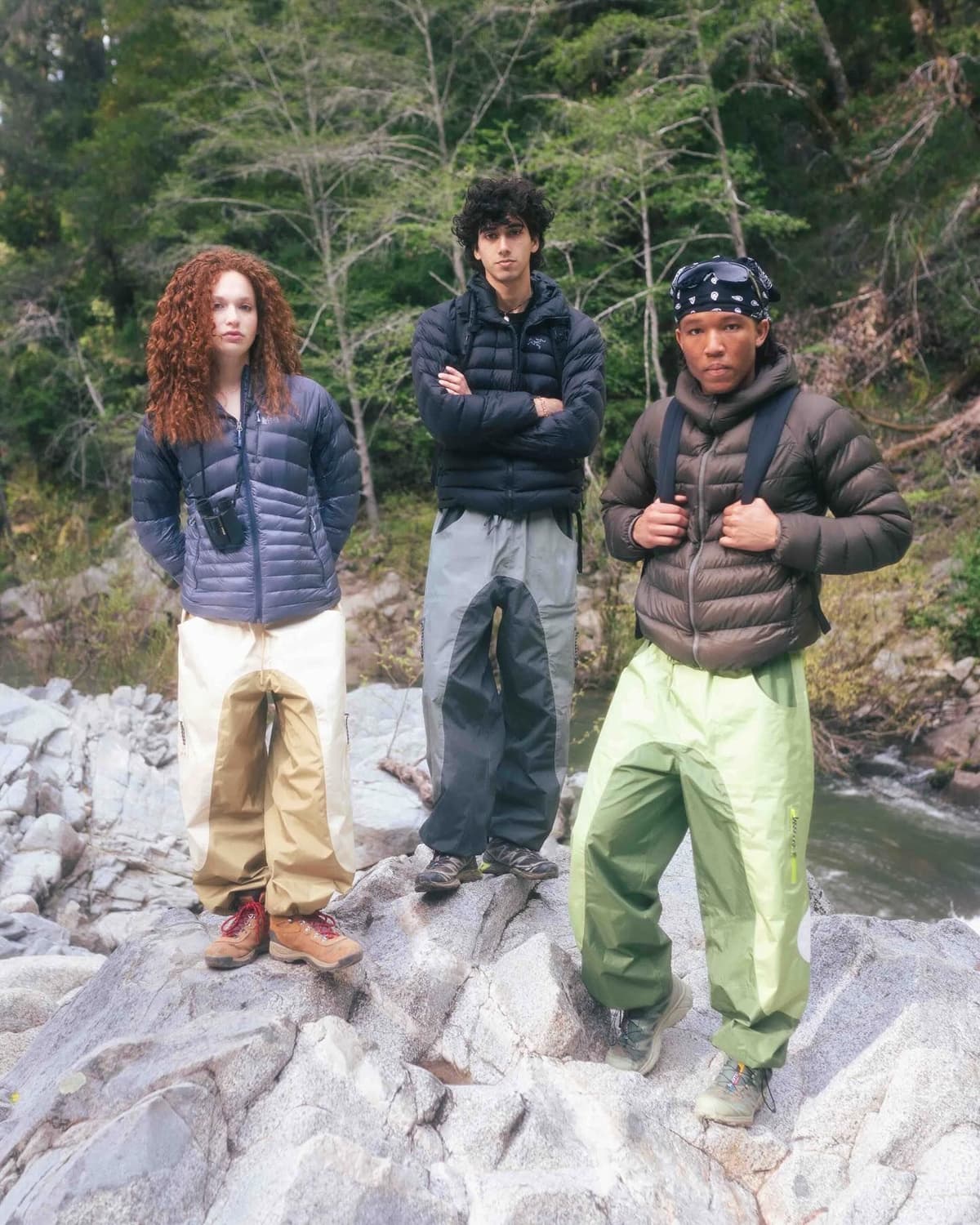 Three people wearing puffer jackets and distinct two-tone outdoor pants pose on rocks by a river.