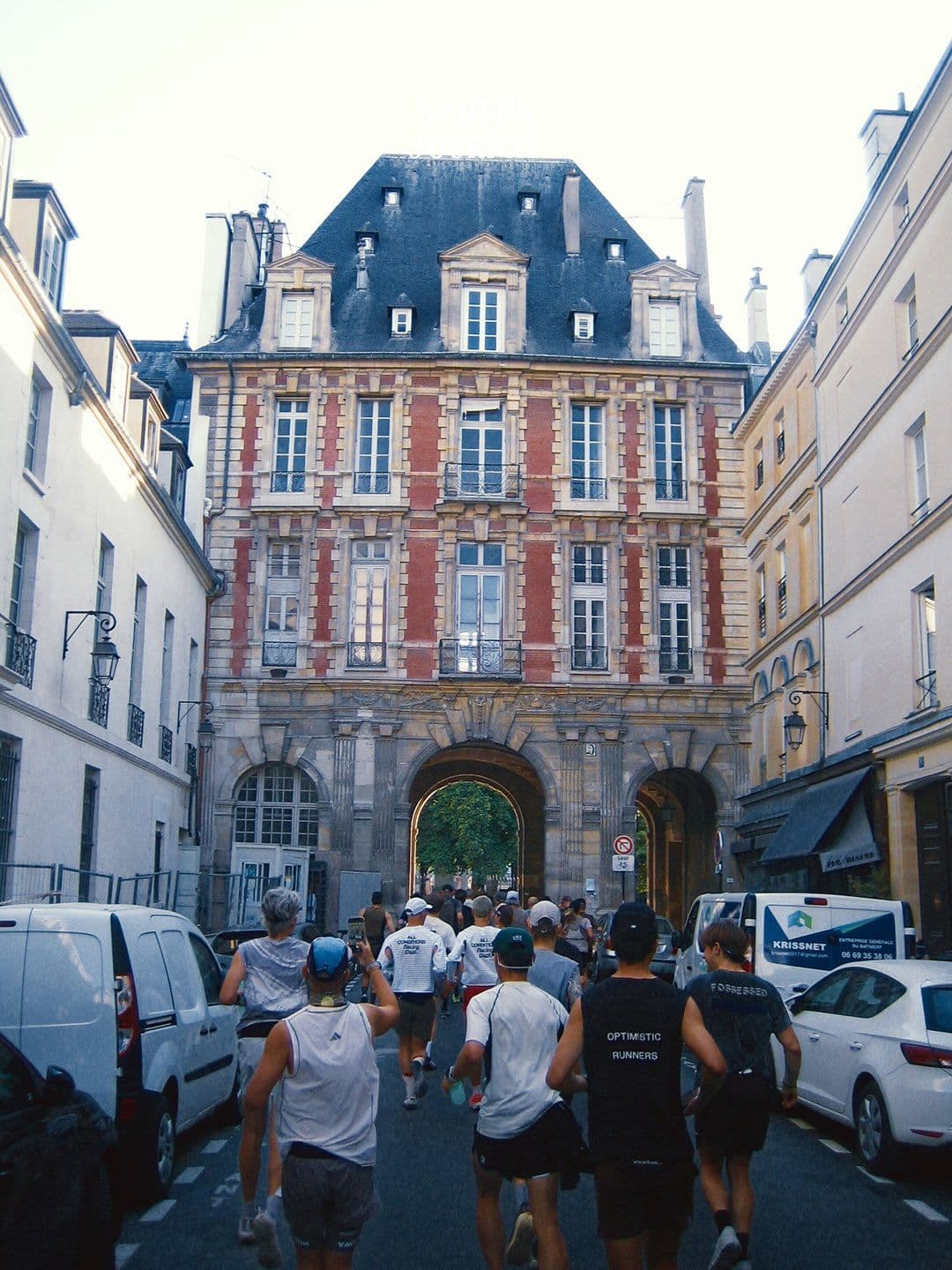 A group of runners moves down a narrow street towards a historic red brick building with a large archway.