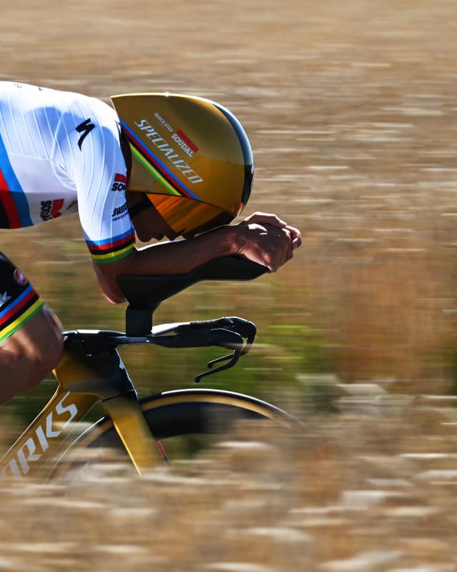 A cyclist in a gold helmet and white rainbow-striped jersey rides a time trial bike, with motion blur in the background.