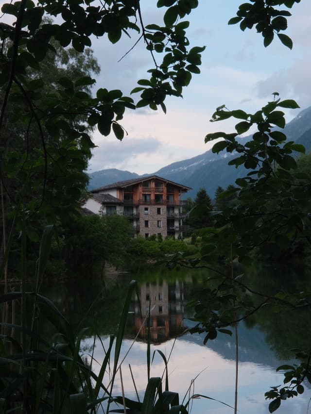 A multistory house with stone and wood details across a tranquil lake reflecting the sky, framed by dark foliage, with mountains in the background.