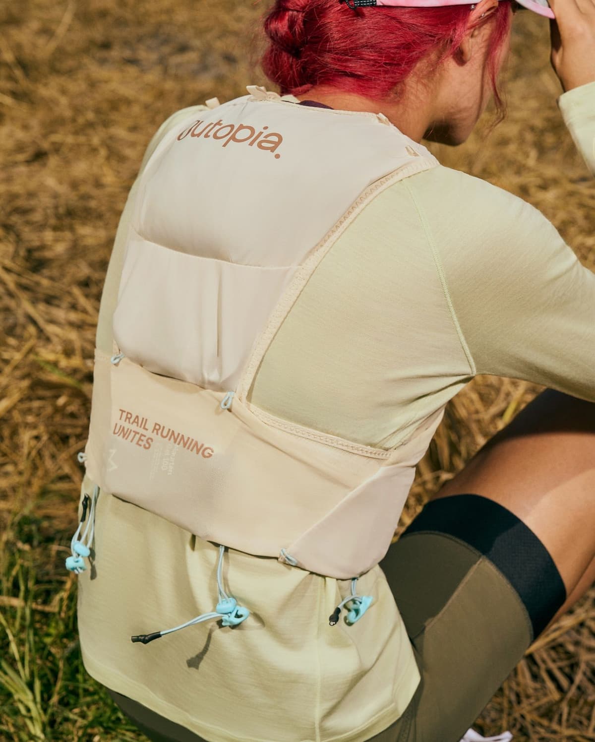 Back view of a person with red hair wearing a beige trail running vest with "Jutopia" and "TRAIL RUNNING UNITES" text, over a light green shirt, kneeling in dry grass.
