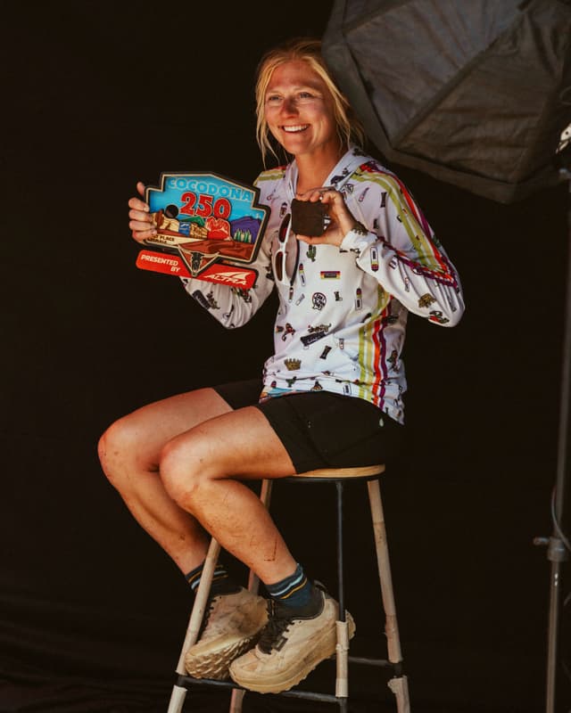 A smiling blonde woman with dirty legs sits, holding a "Cocodona 250" award and a small snack.