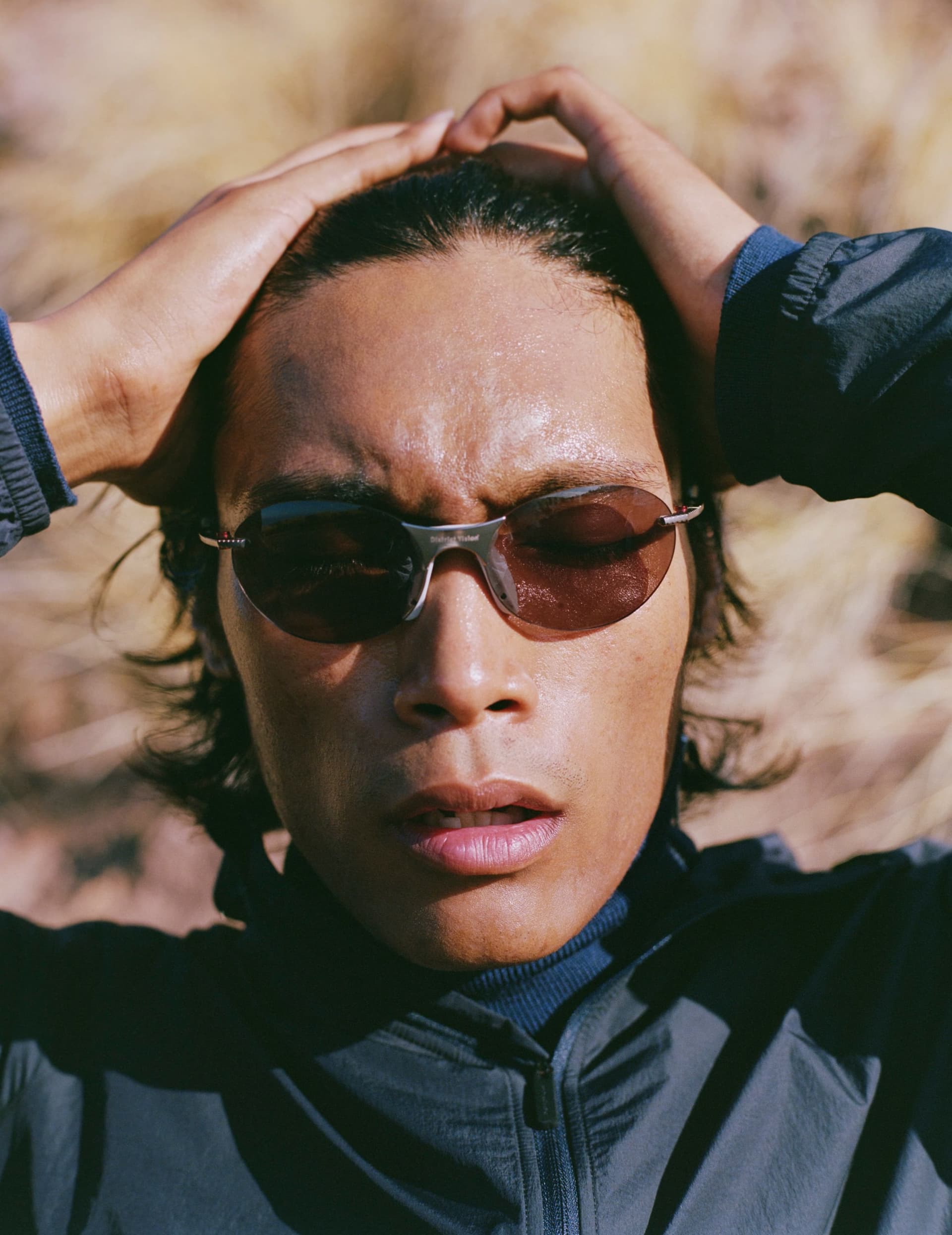 A person wearing sunglasses and a dark jacket, with hands clasped behind their head, looks up.