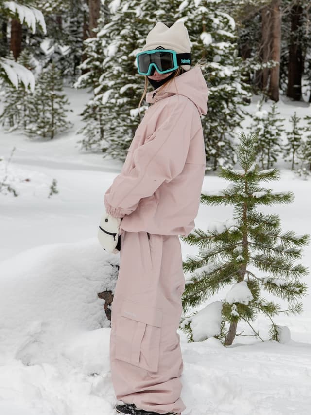 A person in a pale pink snowsuit, cream beanie, and teal ski goggles stands in a snowy forest.