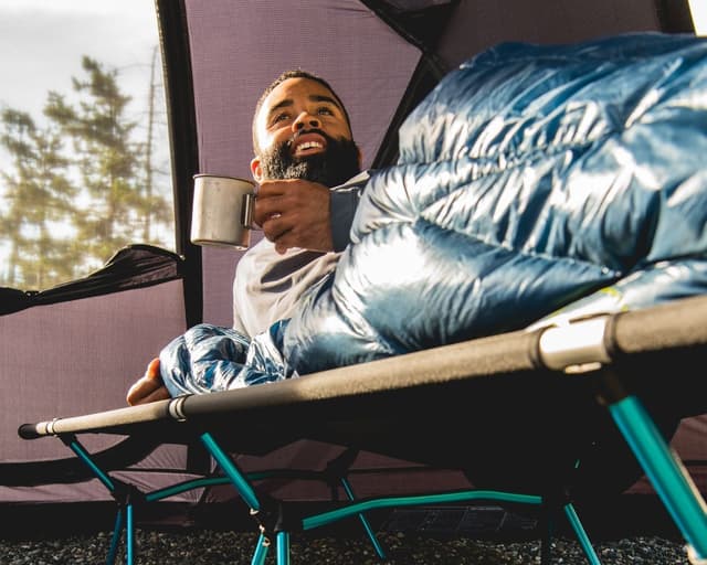 A bearded man lies on a camping cot inside a tent, holding a mug and looking up contentedly.