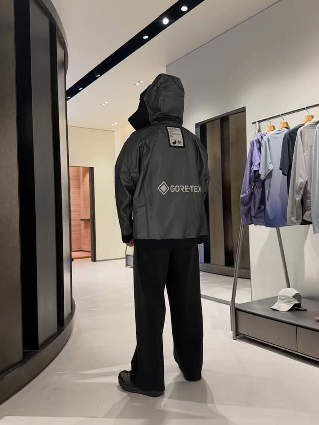 A person from behind wearing a gray hooded Gore-Tex jacket and black pants in a modern store.