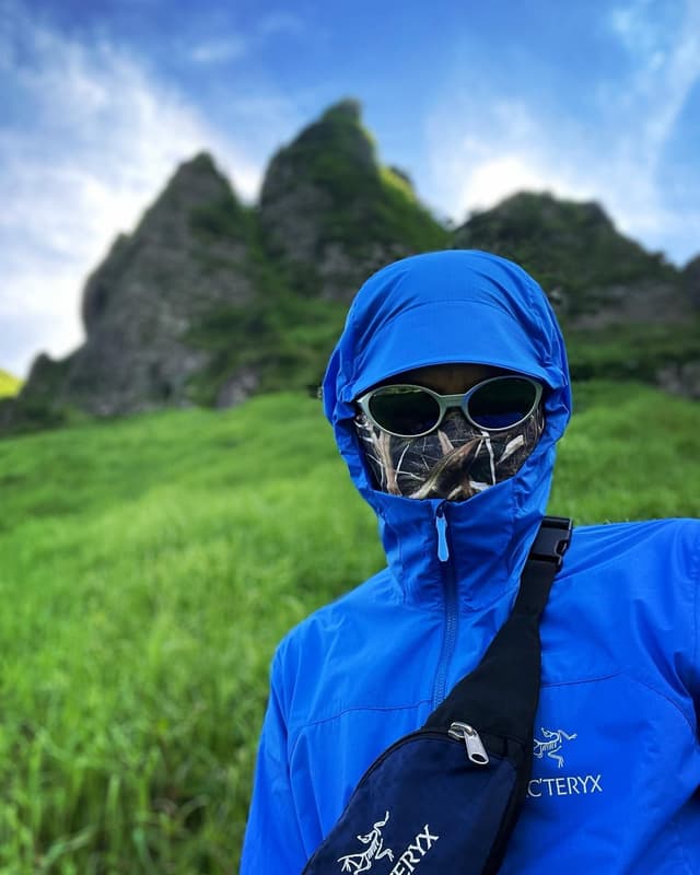 A person in a blue hooded jacket, sunglasses, and camouflage face covering, wearing an Arc'teryx fanny pack, stands before a large green mountain.
