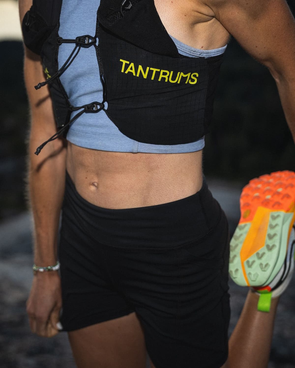 A person wearing a black running vest with "TANTRUMS" logo and black shorts stretches, holding their foot up to show the bright orange sole of their shoe.