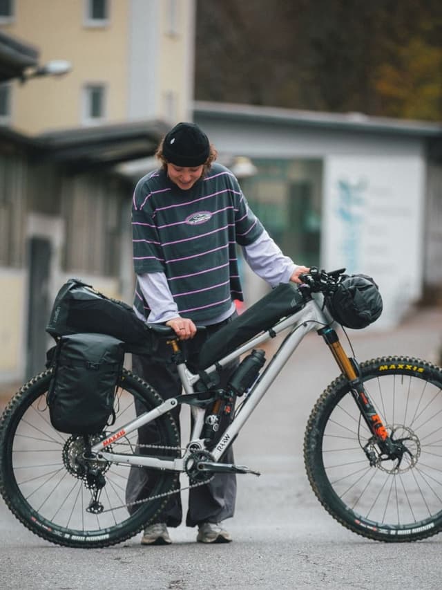 A person with a beanie smiles while standing next to a bikepacking mountain bike loaded with bags.